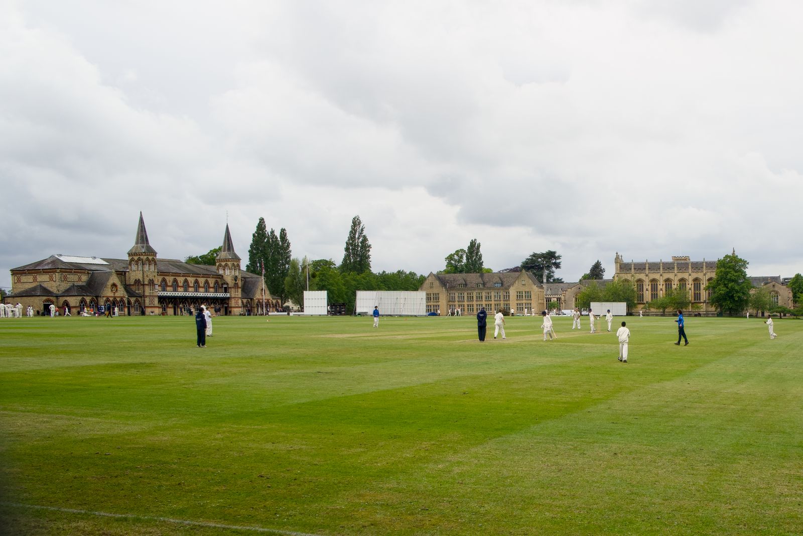 Outdoor cricket match on a green field with a historic school building backdrop